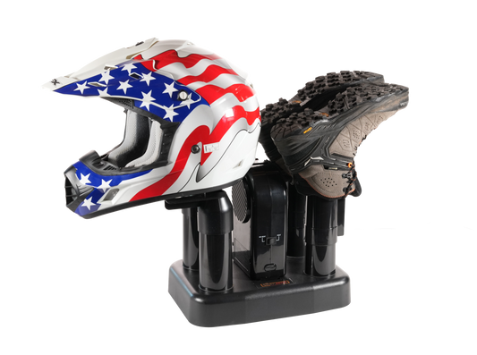 A black helmet and shoe dryer holding a black athletic shoe, with a white motocross helmet featuring a red, white, and blue American flag star pattern resting on the left side, isolated on a white background.
